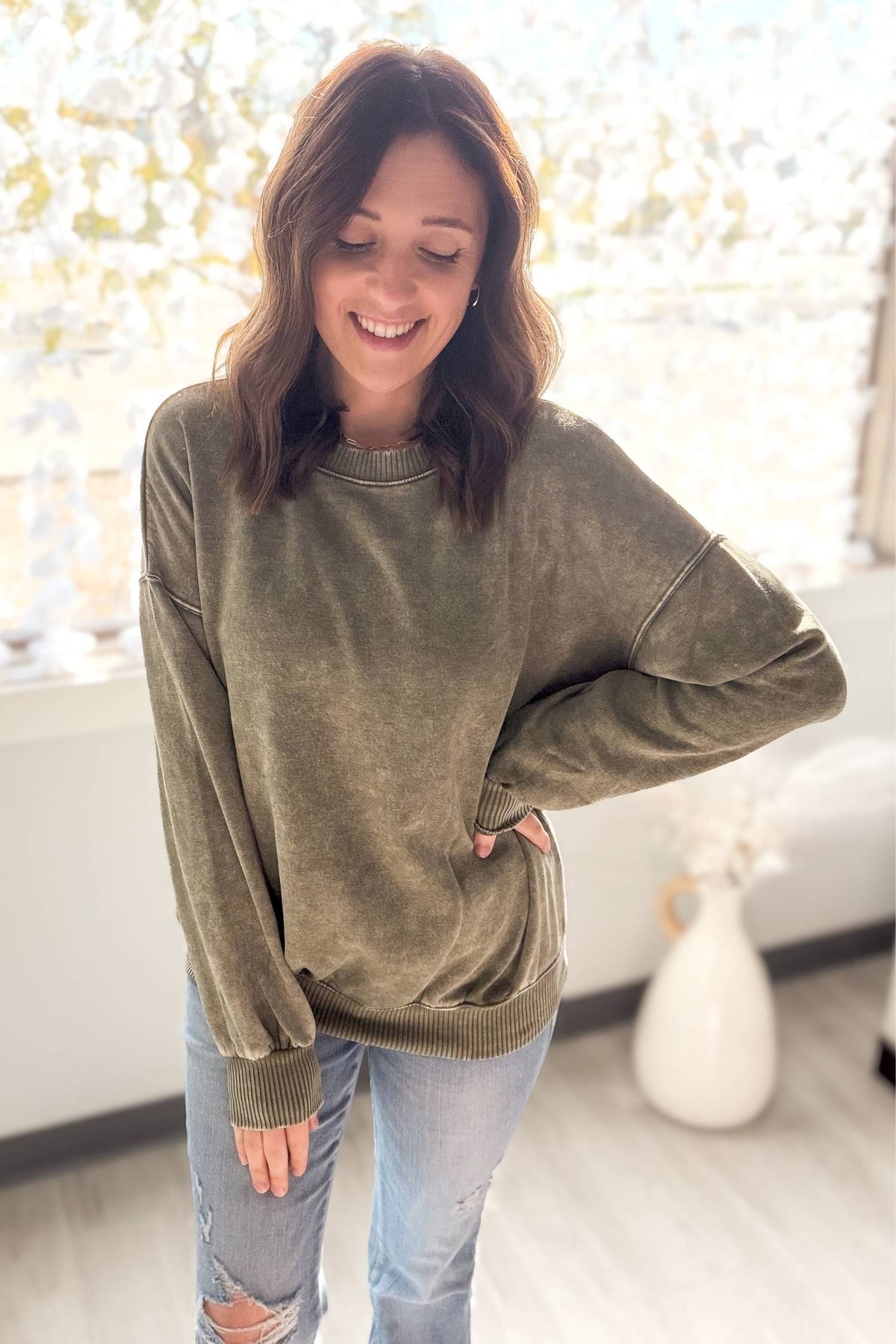 Woman wearing a olive sweater and jeans standing indoors with a blurred background