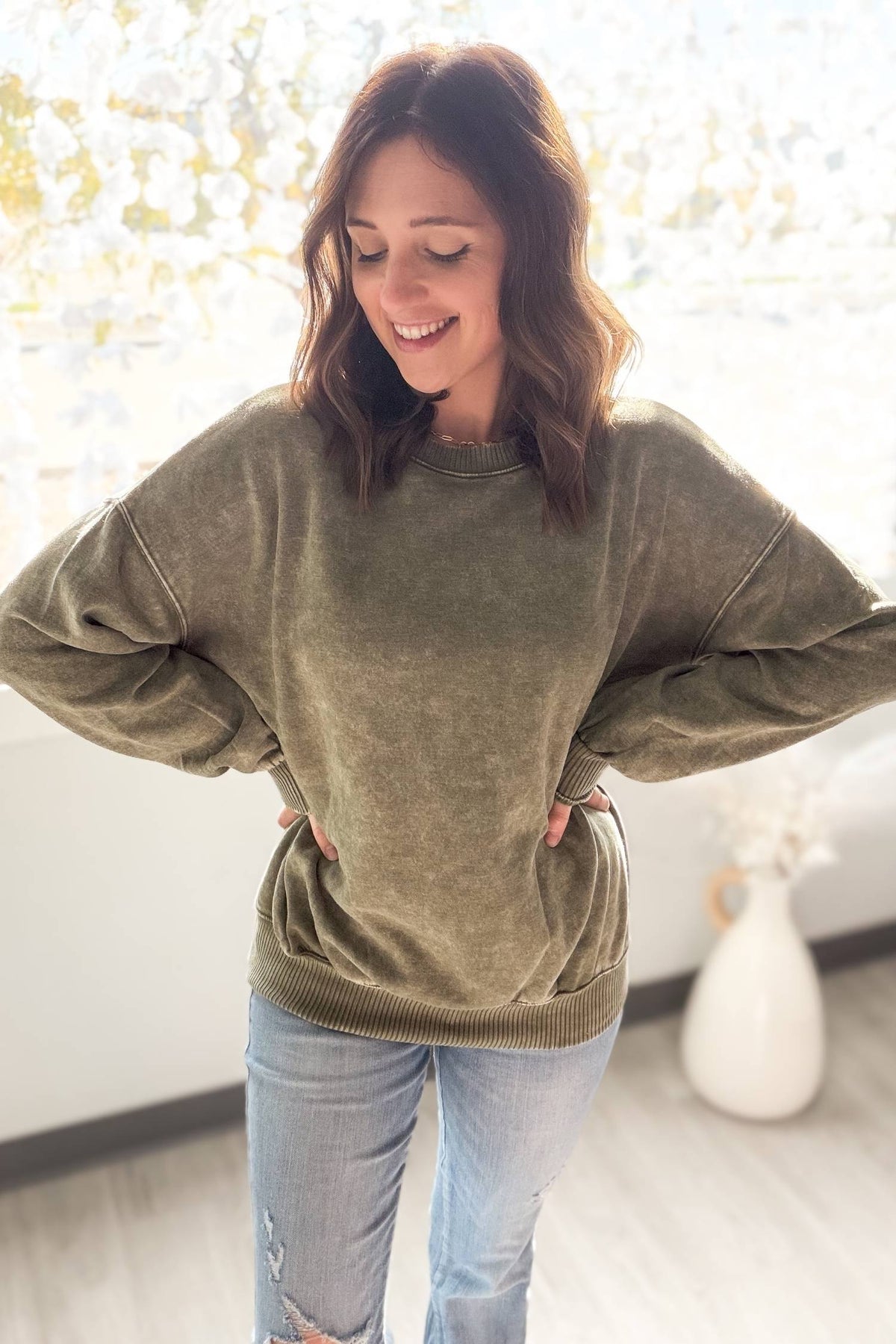 Woman wearing a olive sweater and light blue jeans indoors.
