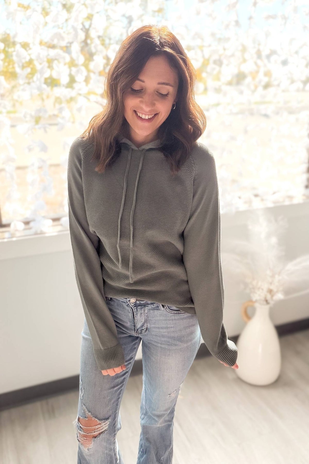 Woman wearing a dusty jade hoodie and ripped jeans indoors with a blurred background
