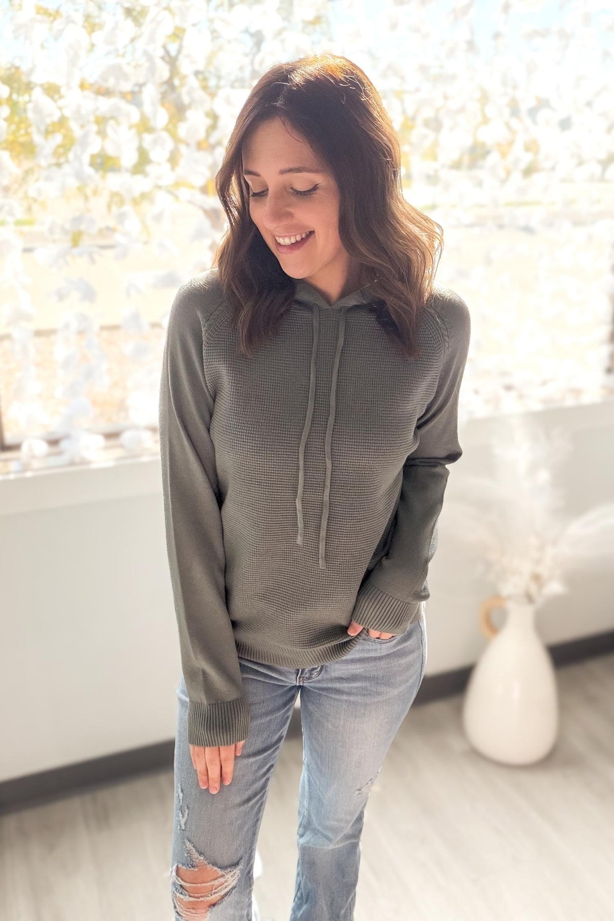 Woman wearing a dusty jade hoodie and ripped jeans indoors with a blurred background