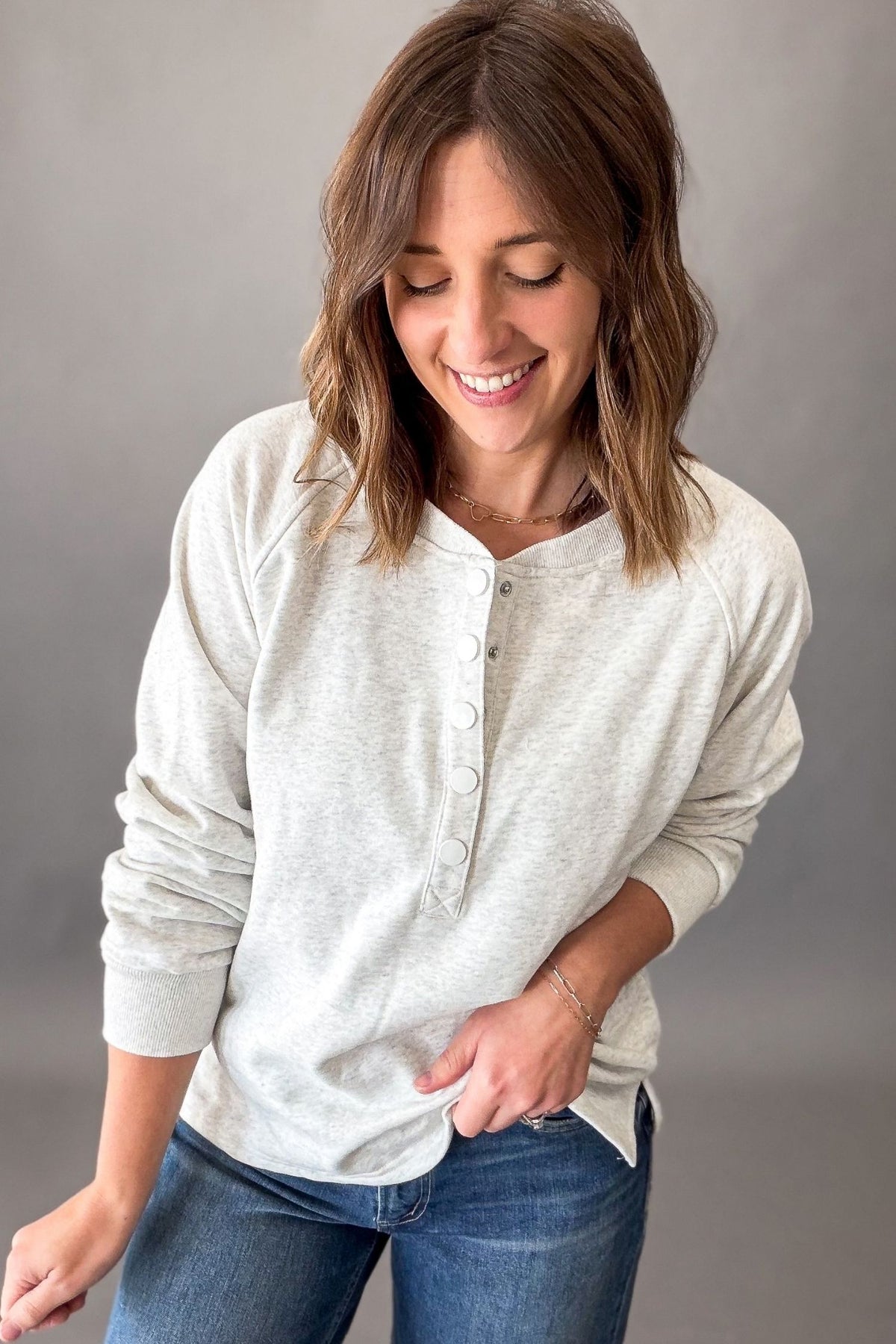 A woman wearing a light heather grey long-sleeve henley shirt with blue jeans, standing against a neutral gray studio background.