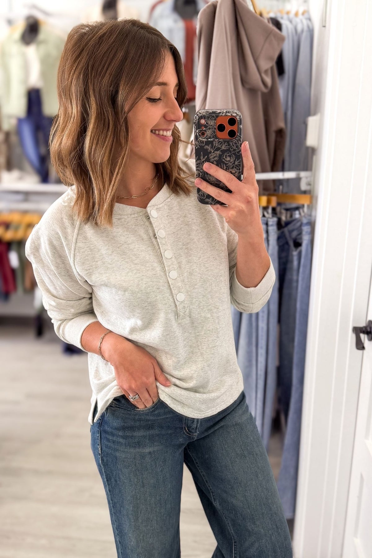 A woman takes a mirror selfie in a clothing store, wearing a light beige heathered henley shirt and blue jeans.
