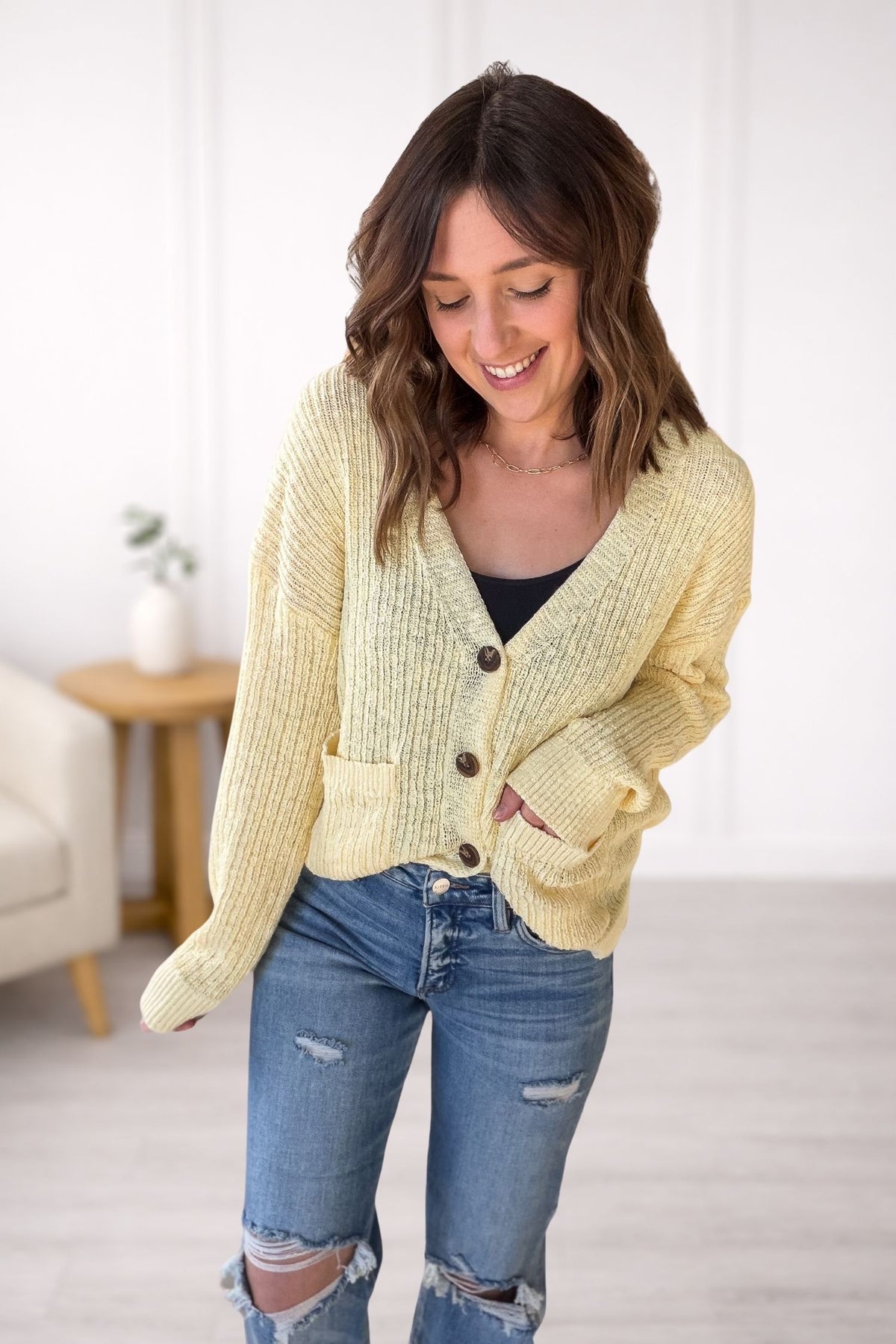 Woman wearing a yellow cardigan and blue jeans in a room with a white wall and a small table.