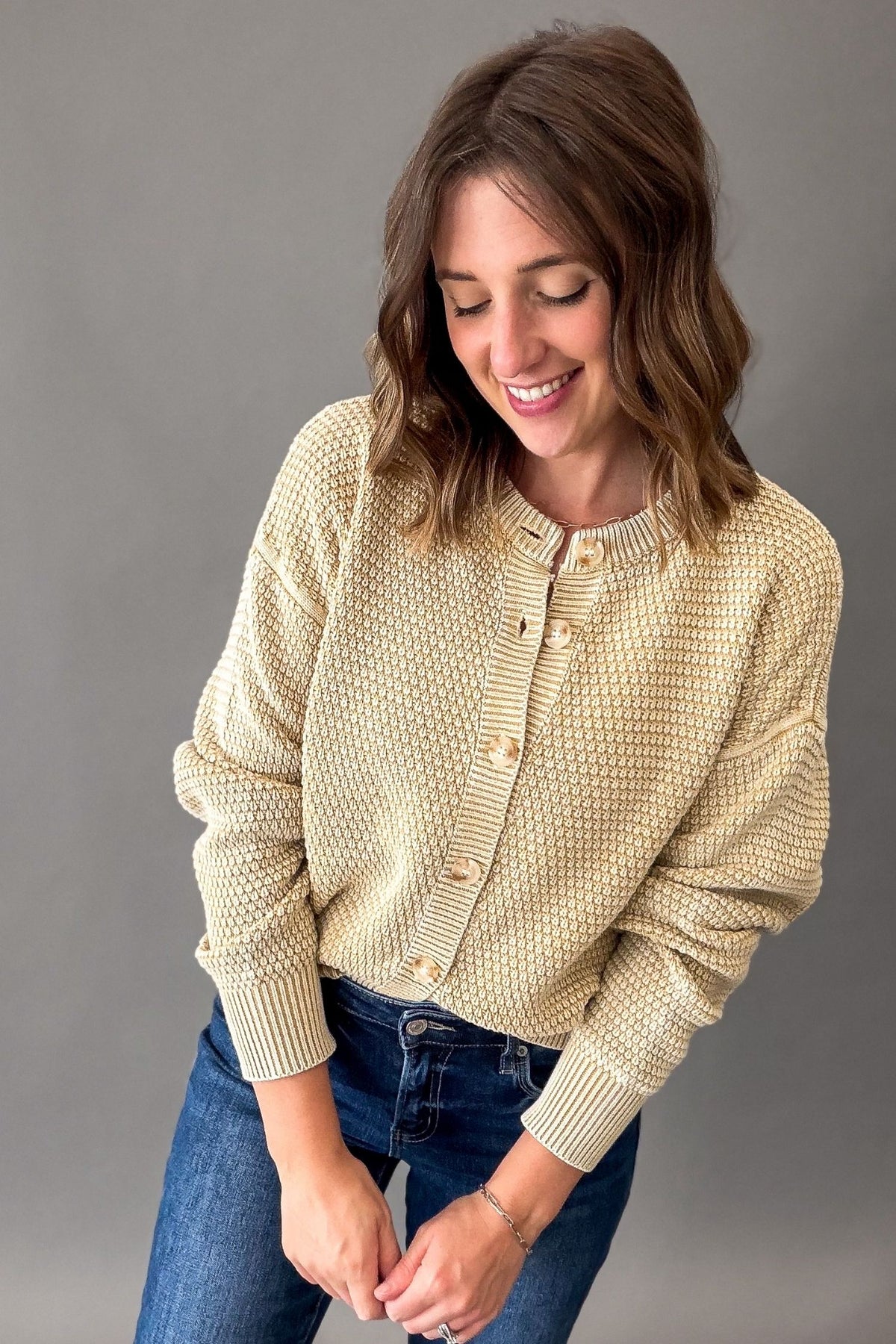 A woman wearing a textured beige-colored knit cardigan with buttons and blue jeans, standing against a solid gray studio background.