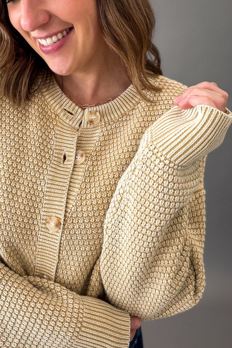 A close-up of a woman wearing a textured beige knit cardigan sweater with a button front, shown against a neutral gray background.