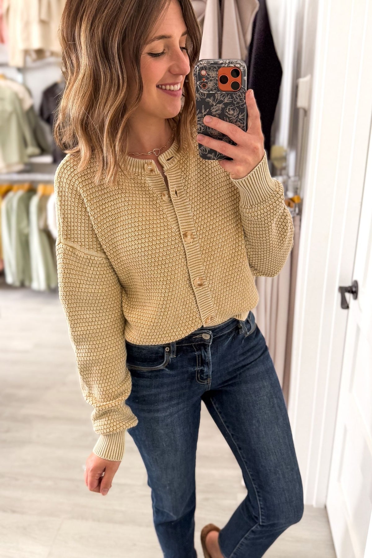 A woman takes a mirror selfie in a boutique while wearing a textured, waffle-knit cropped cardigan in a warm beige color, paired with dark blue jeans.