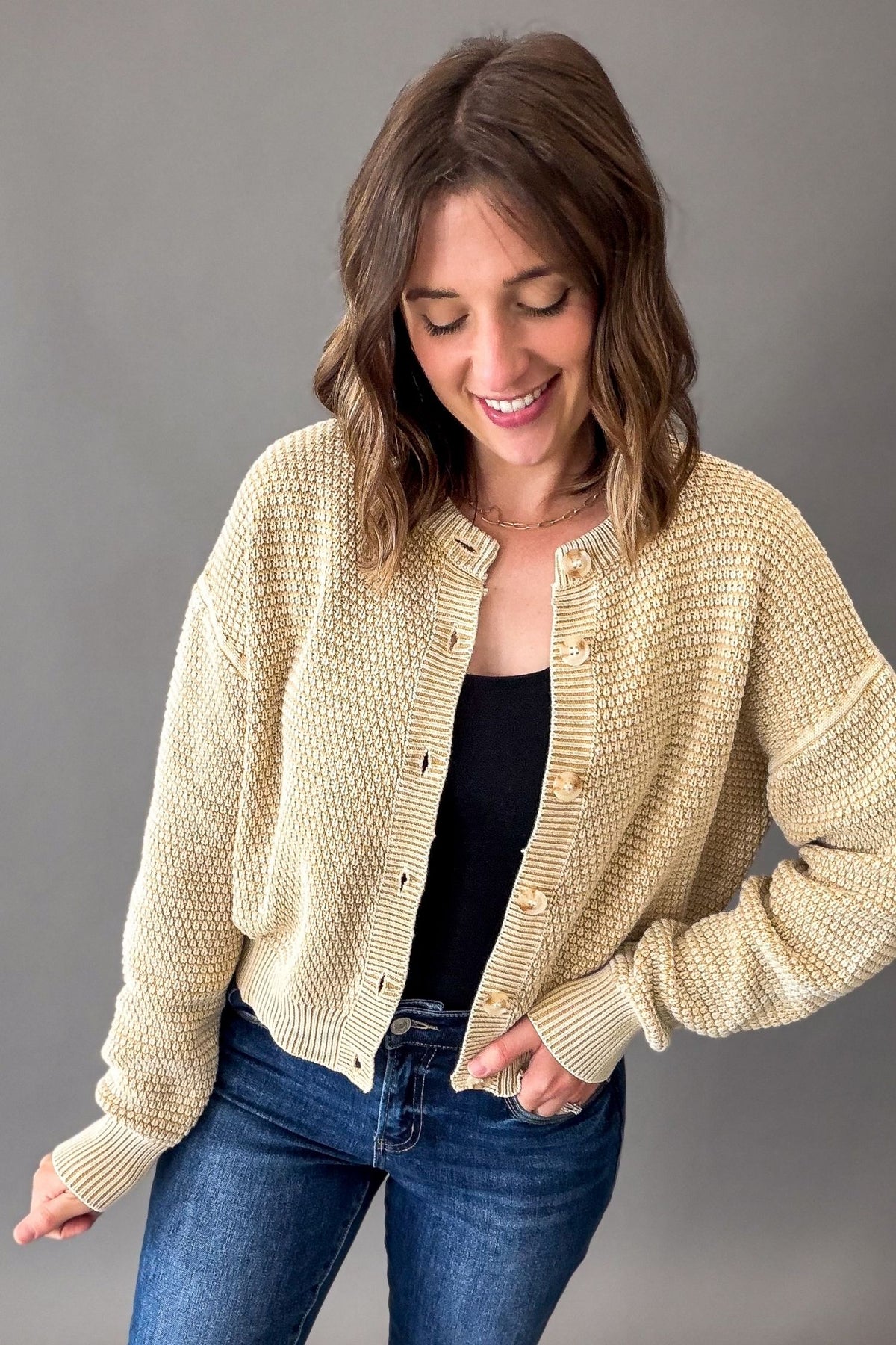 A woman wearing a beige-colored waffle-knit button-up cardigan over a black top and blue jeans, standing against a solid gray studio background.