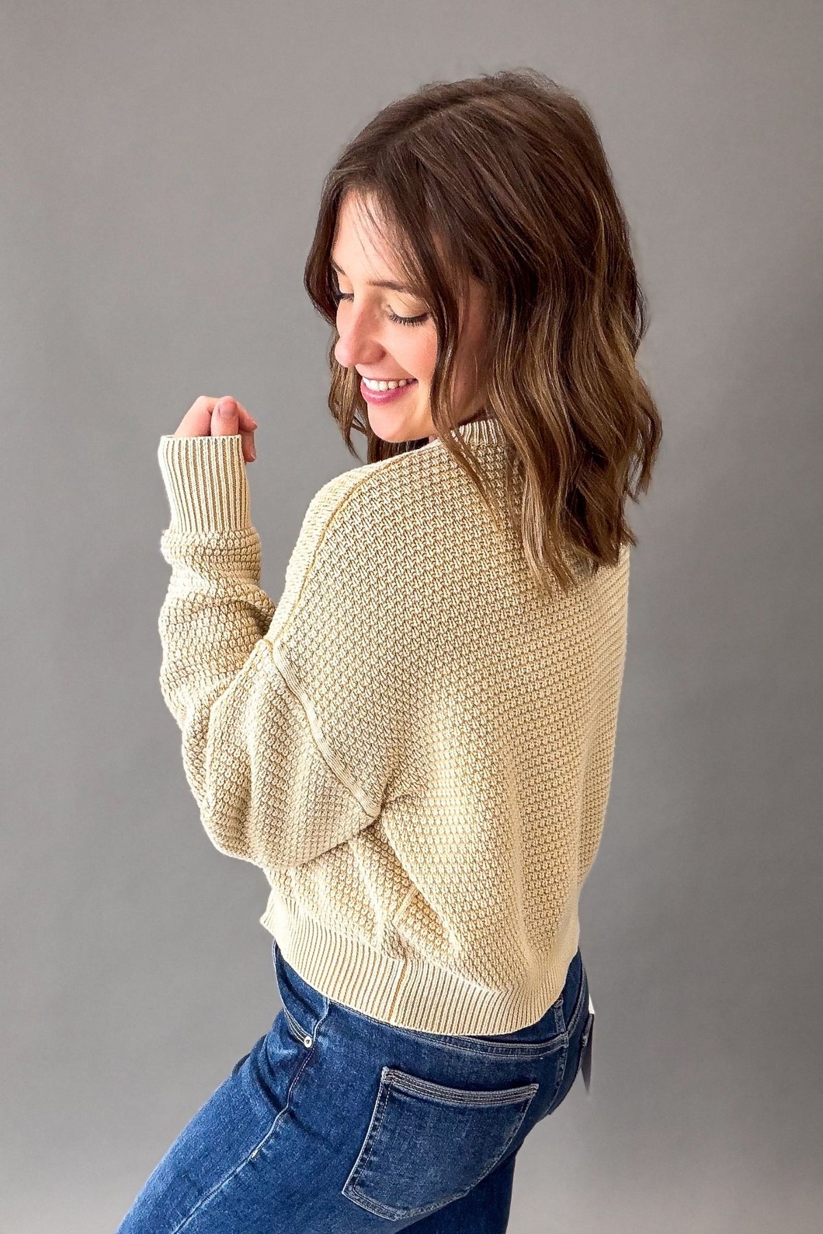 A woman with shoulder-length brown hair is shown from the back wearing a beige-colored textured knit sweater and blue jeans against a solid gray studio background.