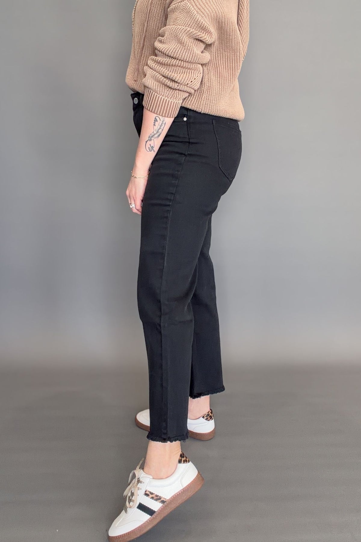 Side view of black high rise crop bootcut jeans with frayed hem and slight flare, styled with white sneakers.