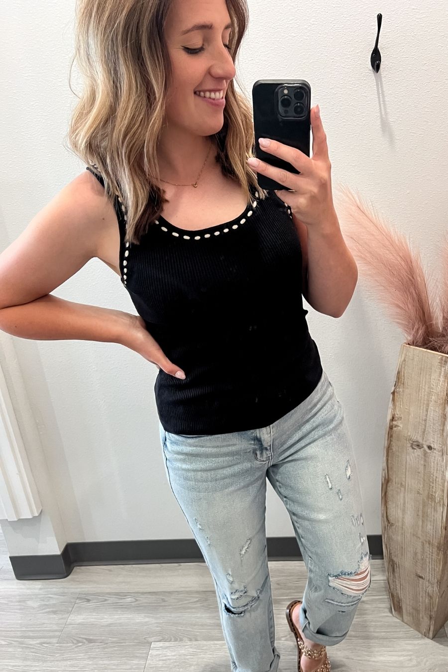 Woman taking a mirror selfie wearing a black tank top with white stitching and light blue jeans.