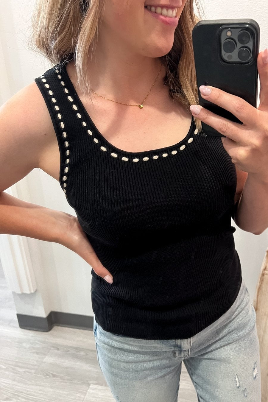 Woman taking a mirror selfie wearing a black tank top with ivory stitching and light blue jeans.