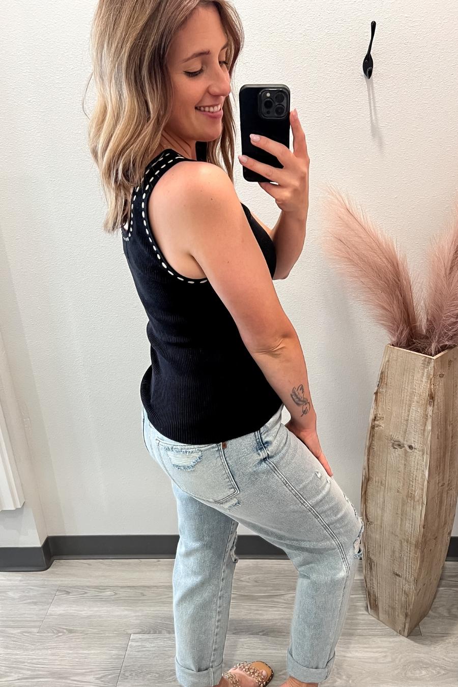 Woman taking a mirror selfie wearing a black sleeveless top and light blue jeans, standing in a room with a decorative plant.