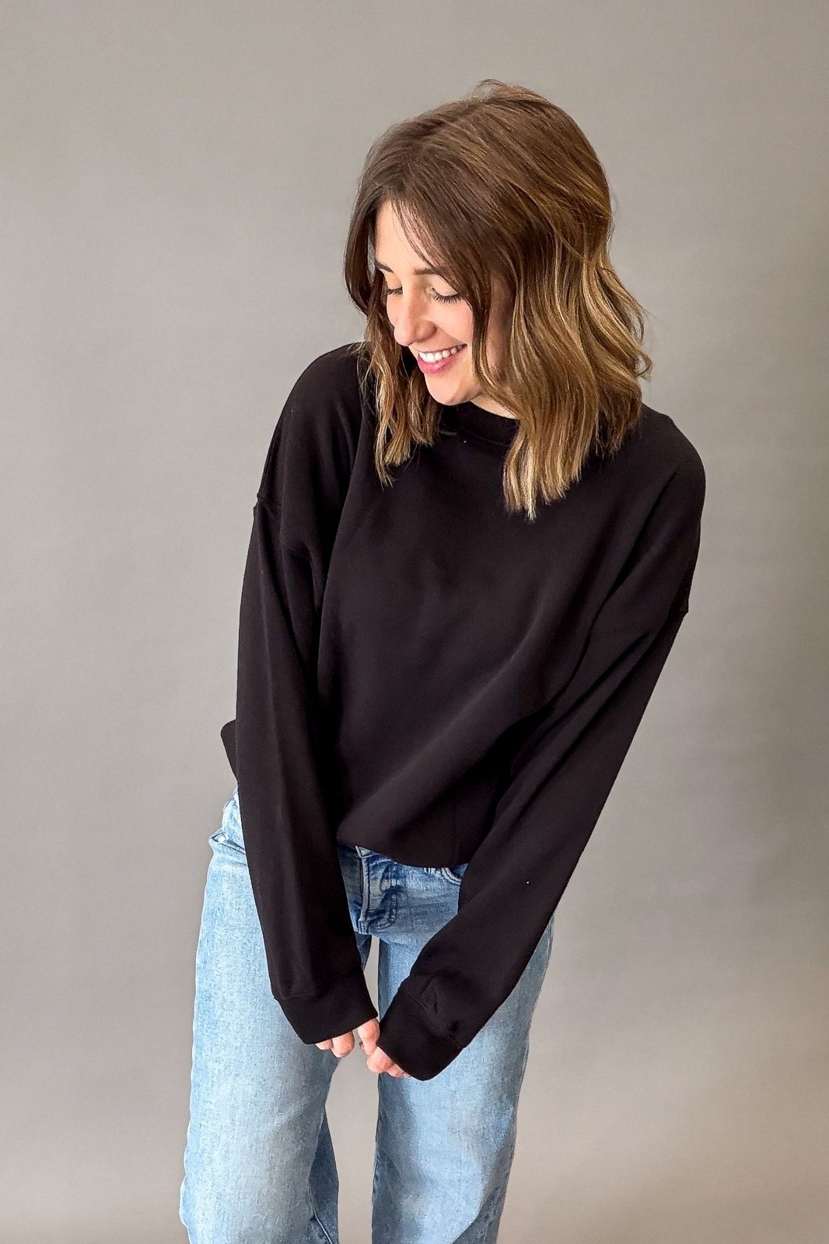 A woman with shoulder-length brown hair wearing a black long-sleeve crewneck sweatshirt and light blue jeans, standing against a neutral gray studio background.