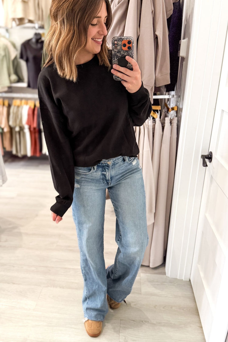 A woman takes a mirror selfie in a clothing boutique, wearing a black long-sleeve crewneck sweater and light-wash blue wide-leg jeans.