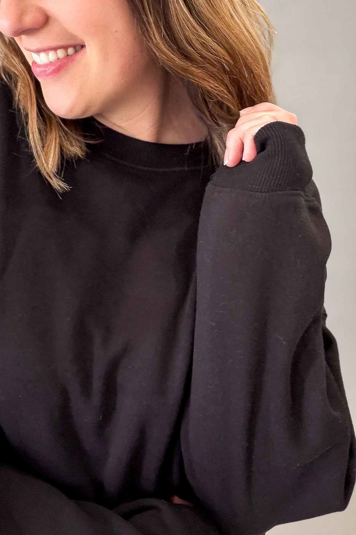A close-up of a person wearing a black crewneck sweatshirt, showing the ribbed collar and sleeve cuffs, with the model smiling and holding the shoulder.