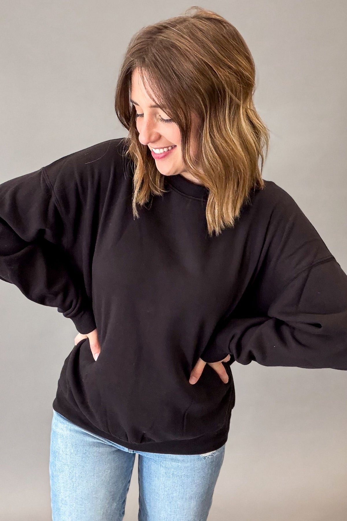 A woman wearing a black oversized crewneck sweatshirt and light blue jeans, standing against a plain gray studio background.