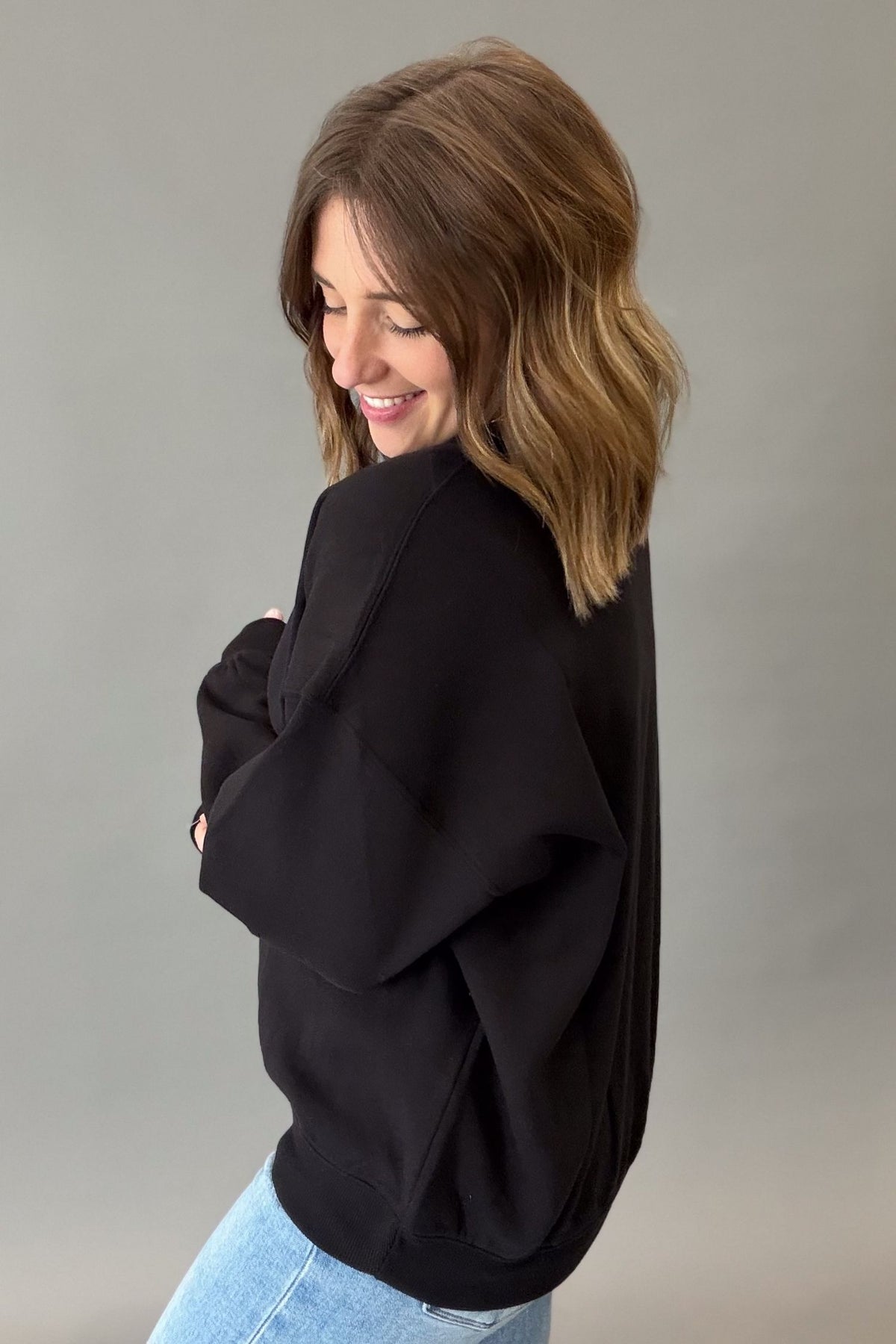 A woman with shoulder-length brown hair is shown from the back wearing a black oversized sweatshirt and light blue jeans against a plain gray studio background.