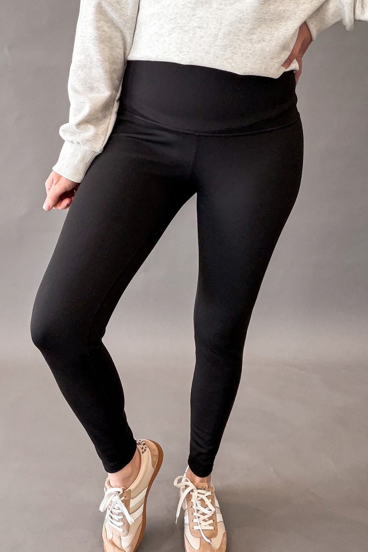 woman wearing black leggings with a heather grey top and sneakers on a grey background.