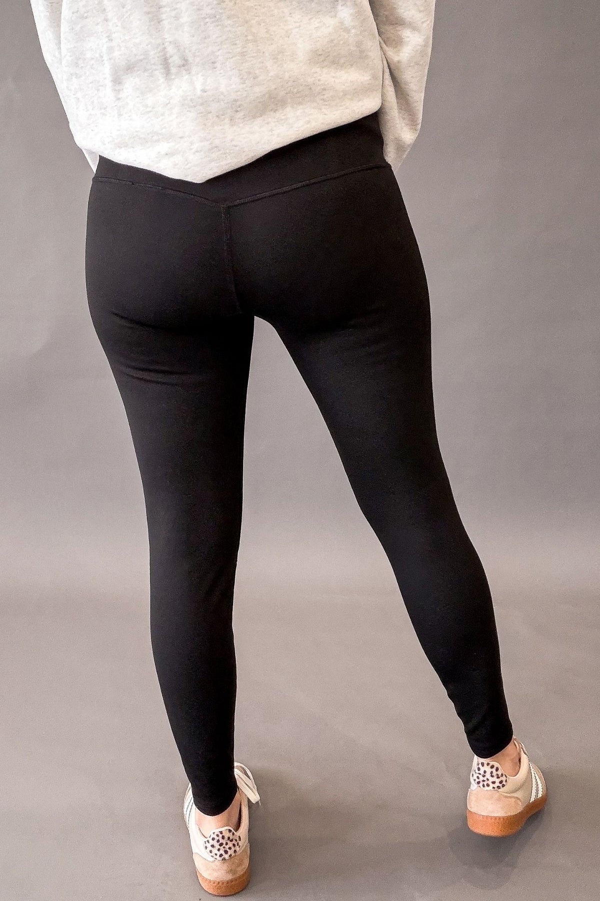 A person shown from the back wearing black leggings, a light gray sweatshirt, and beige sneakers with leopard print details, standing against a neutral gray studio background.