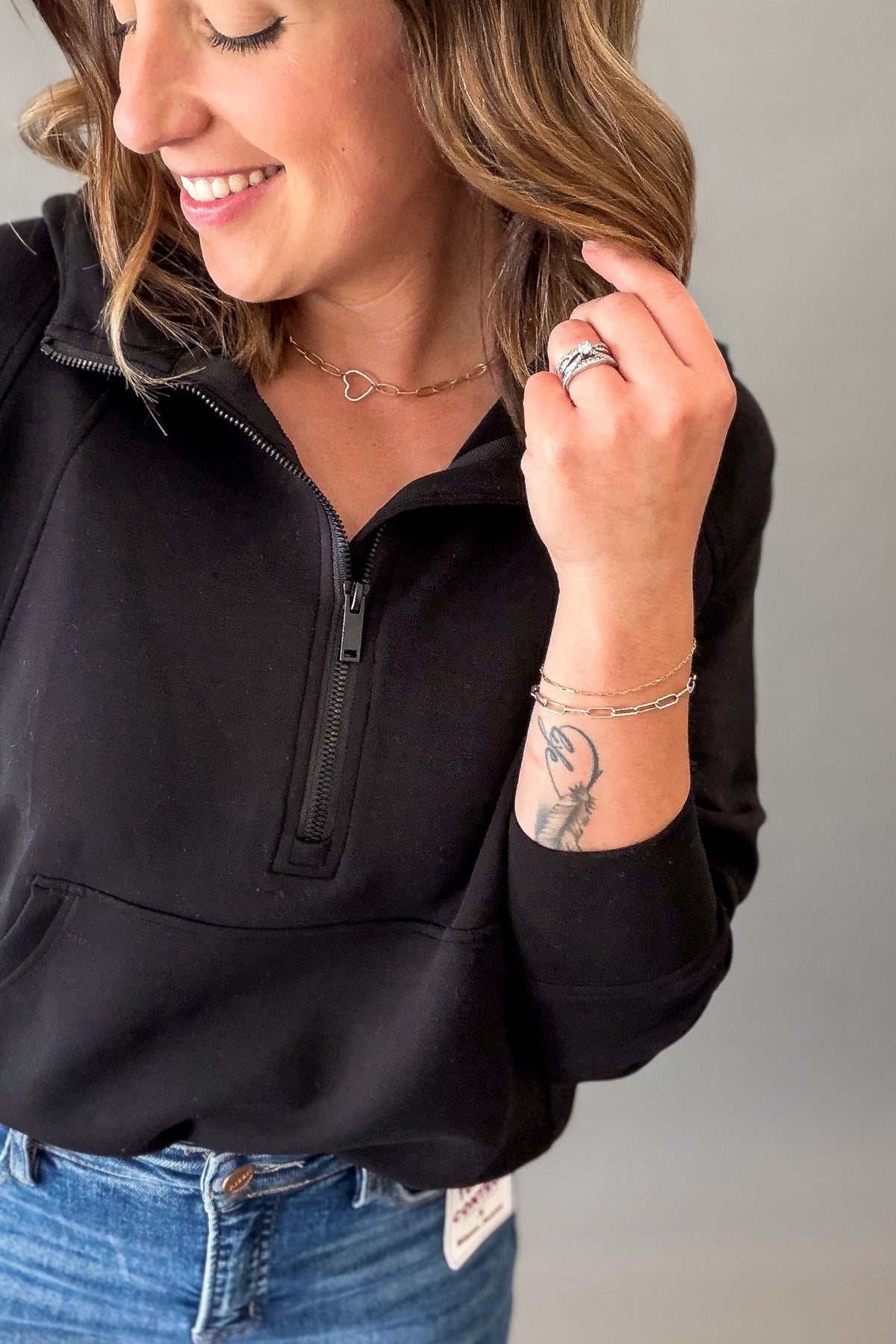 A woman wearing a black quarter-zip pullover sweatshirt and blue jeans, shown from the chest up against a plain gray background.