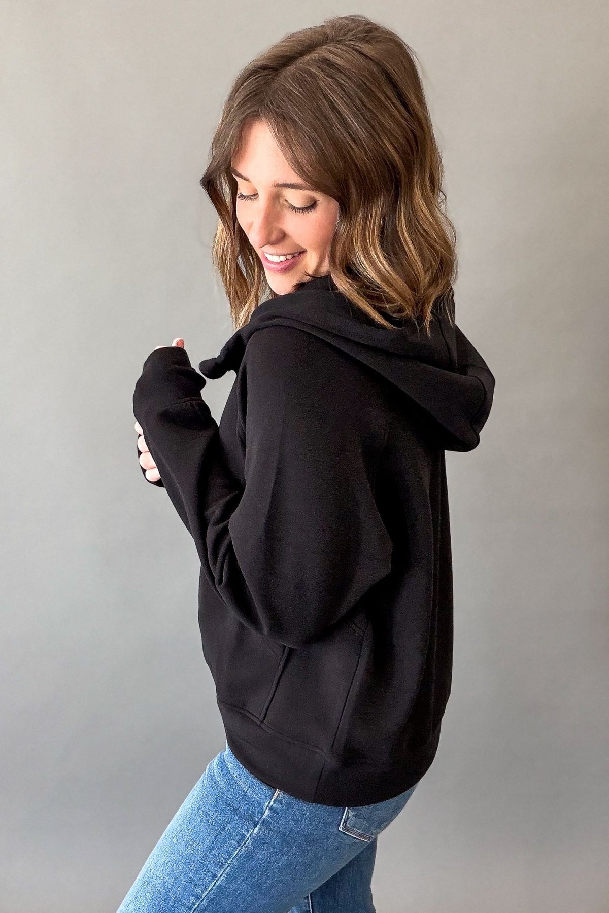 A woman wearing a black pullover hoodie and blue jeans, shown from a side-back angle against a plain gray studio background.