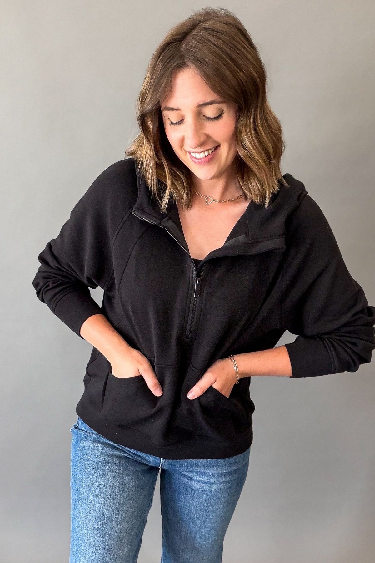 A woman wearing a black half-zip hoodie with a high collar and front pockets, paired with blue jeans, standing against a plain gray background.