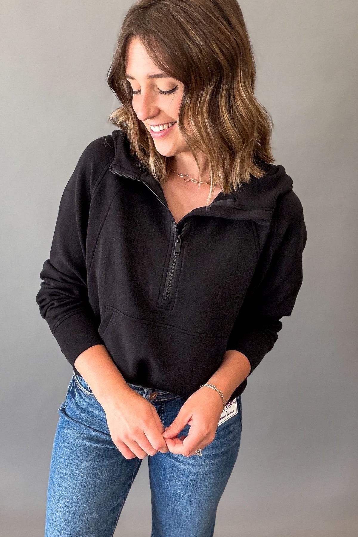 A woman wearing a black half-zip pullover hoodie with a mock neck and drawstring hood, paired with blue jeans, posing against a neutral gray studio background.