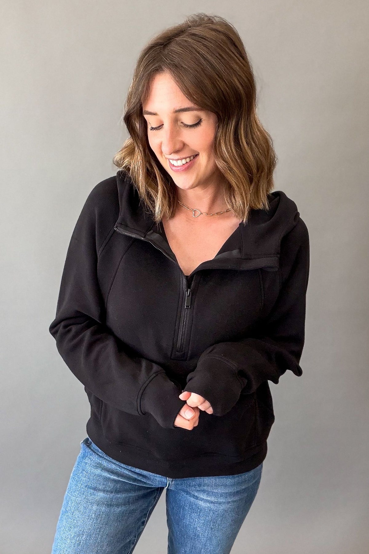 A woman wearing a black half-zip hoodie with a high collar and thumbholes, paired with blue jeans, posing against a solid gray studio background.