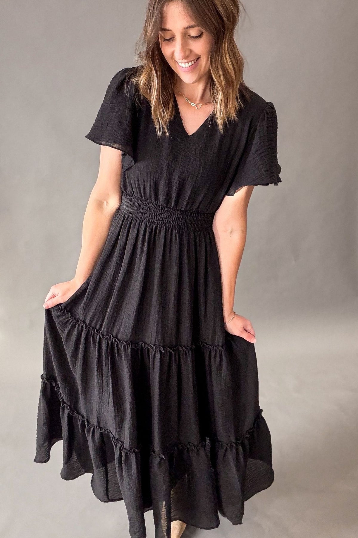 A woman wearing a black tiered midi dress with short flutter sleeves and a smocked waist, standing against a neutral gray studio backdrop.