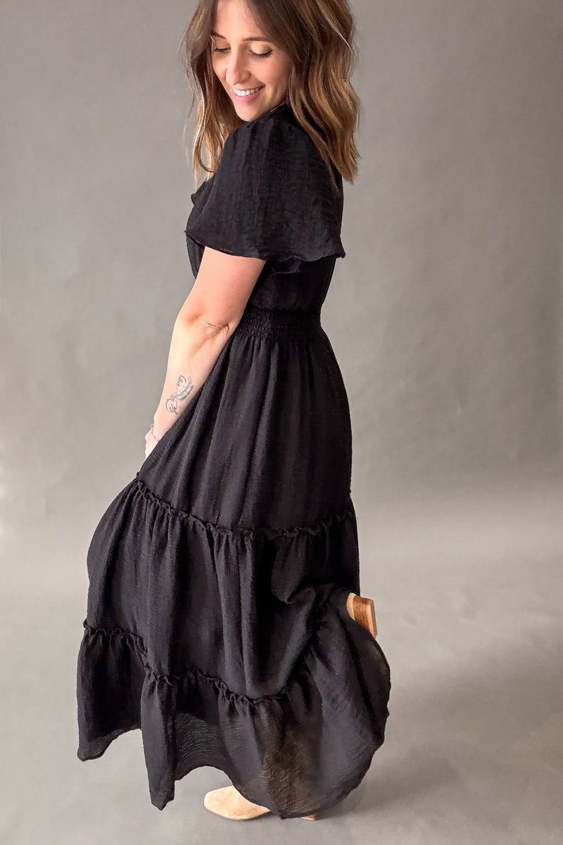 A woman wearing a black tiered maxi dress with short flutter sleeves, shown from a side-back angle against a neutral gray studio background.