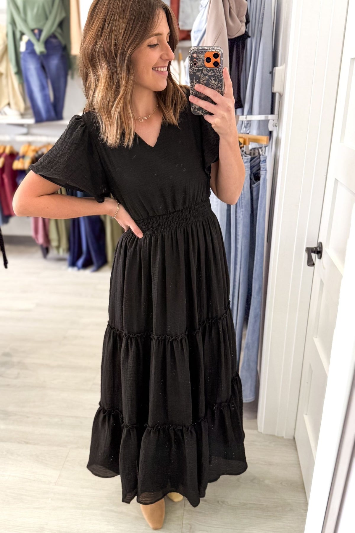 woman taking a selfie in a boutique wearing a black textured maxi dress with flutter sleeves and soft v neckline with a tan colored boot