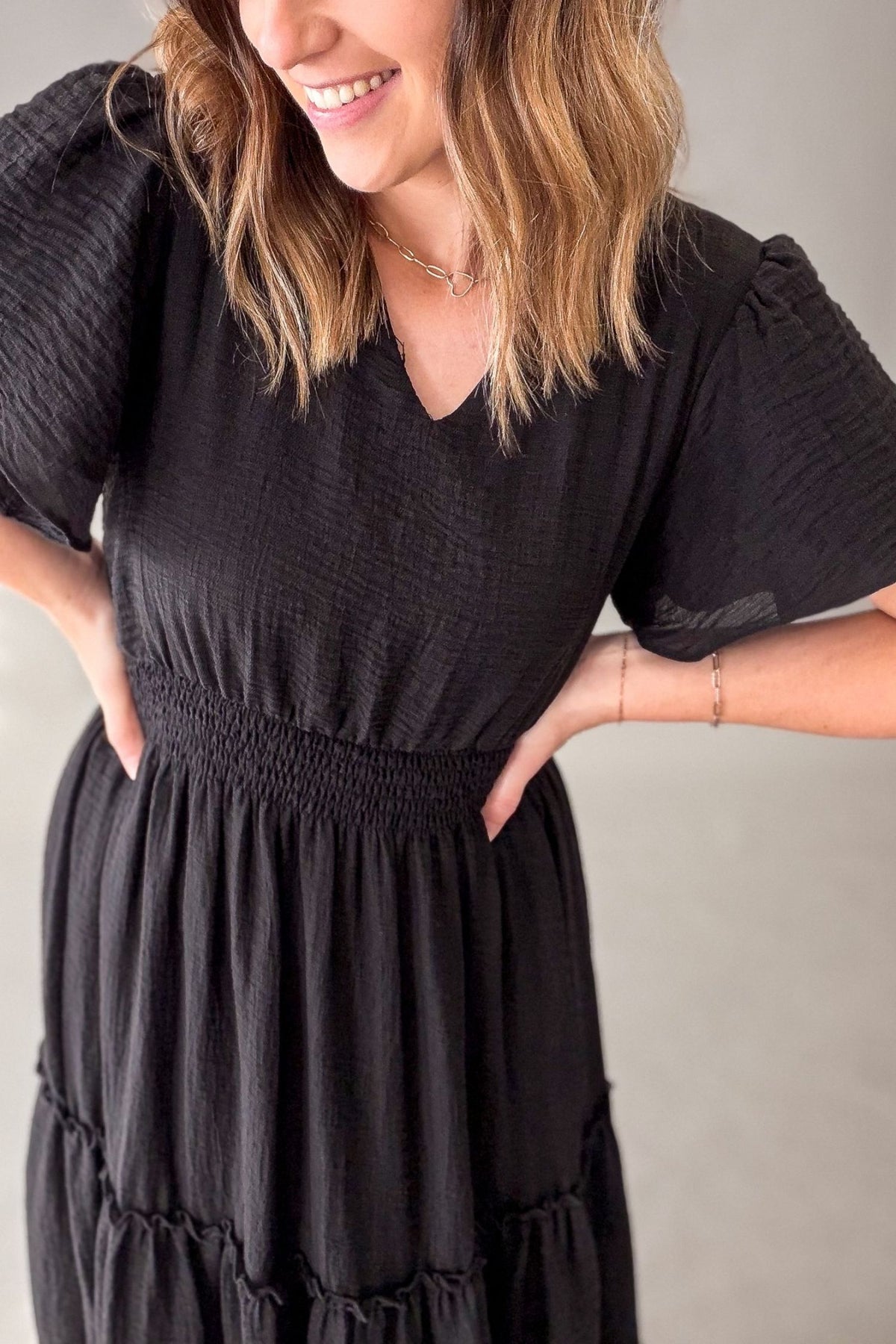 A woman wearing a black textured short-sleeve maxi dress with a smocked waist and tiered skirt, shown from the chest down against a neutral gray background.