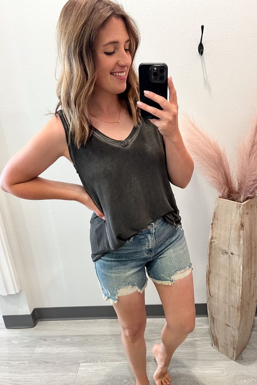 Woman taking a mirror selfie wearing a black tank top and denim shorts, standing in a room with a vase of pink flowers.