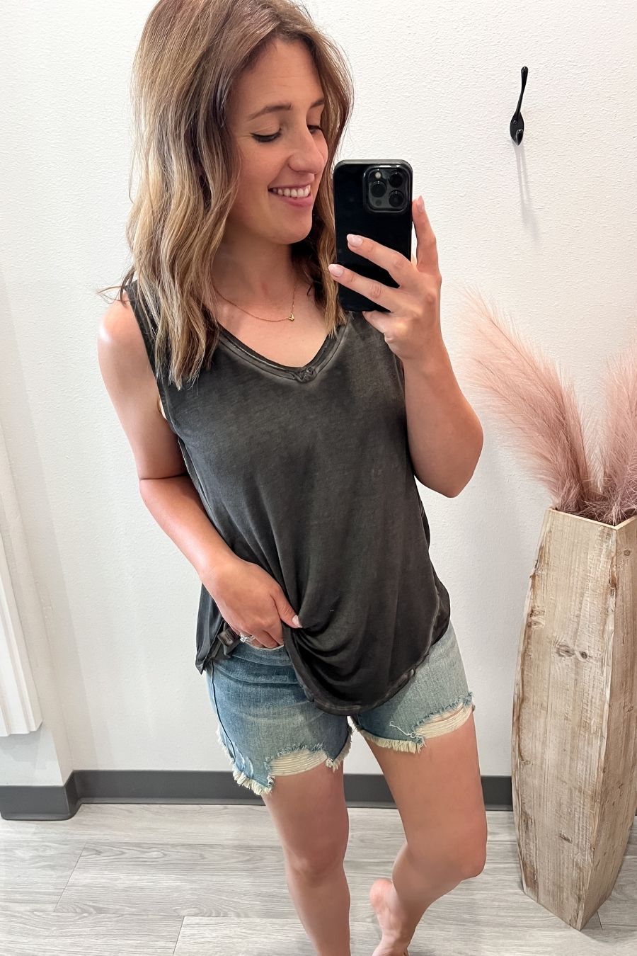 Woman taking a mirror selfie wearing a dark gray tank top and denim shorts, standing in a room with a plant and wooden vase.
