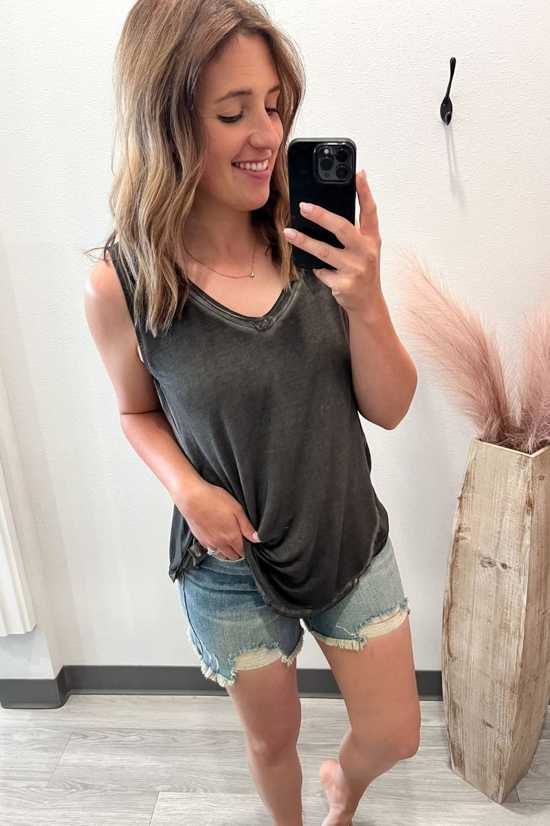 Woman taking a mirror selfie wearing a dark gray tank top and denim shorts, standing in a room with a plant and wooden vase.
