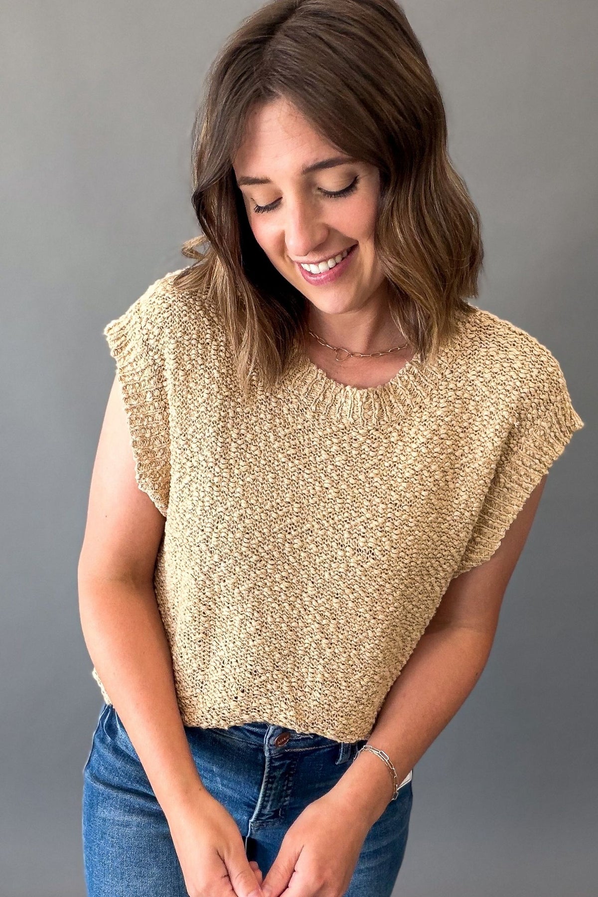 A woman wearing a textured, short-sleeve knit top in a warm beige or light brown color, paired with blue jeans, standing against a plain gray background.