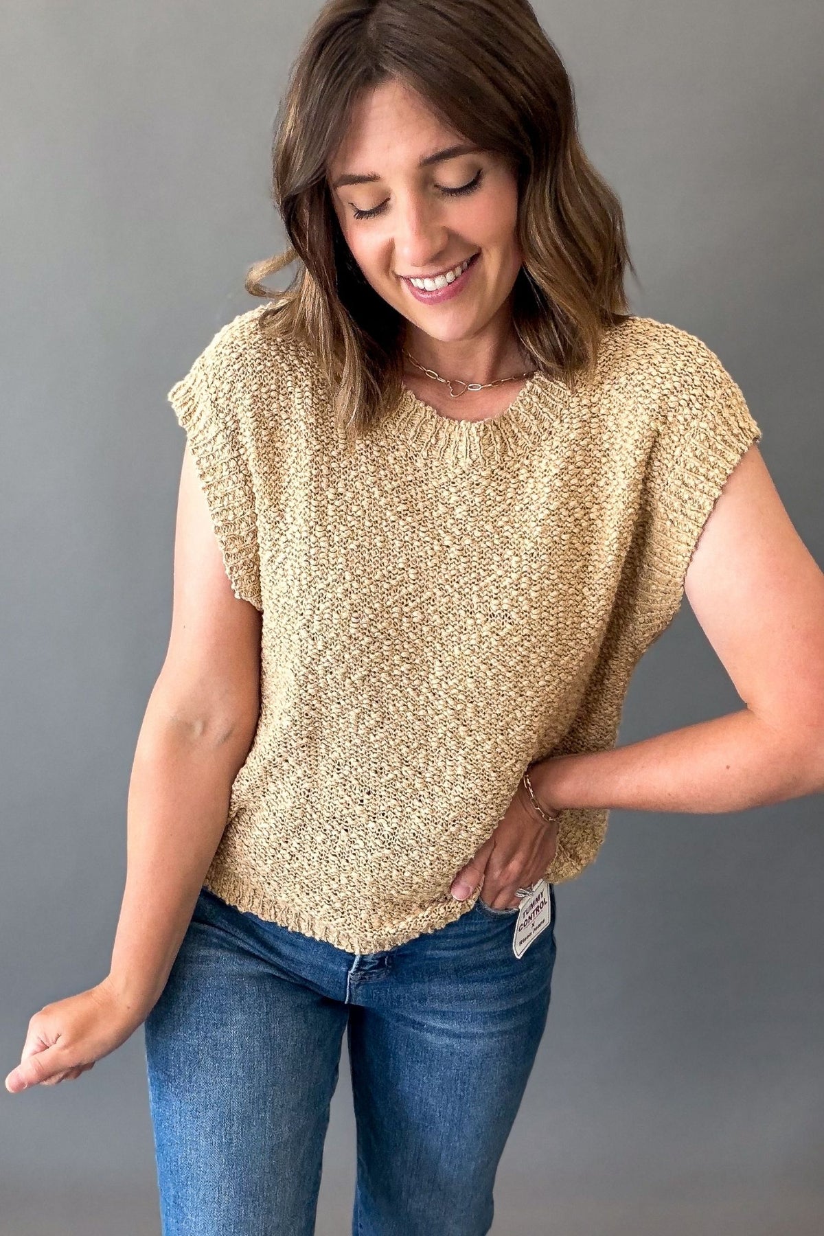 A woman wearing a textured, short-sleeve knit top in a biscuit color, paired with blue jeans, standing against a solid gray background.