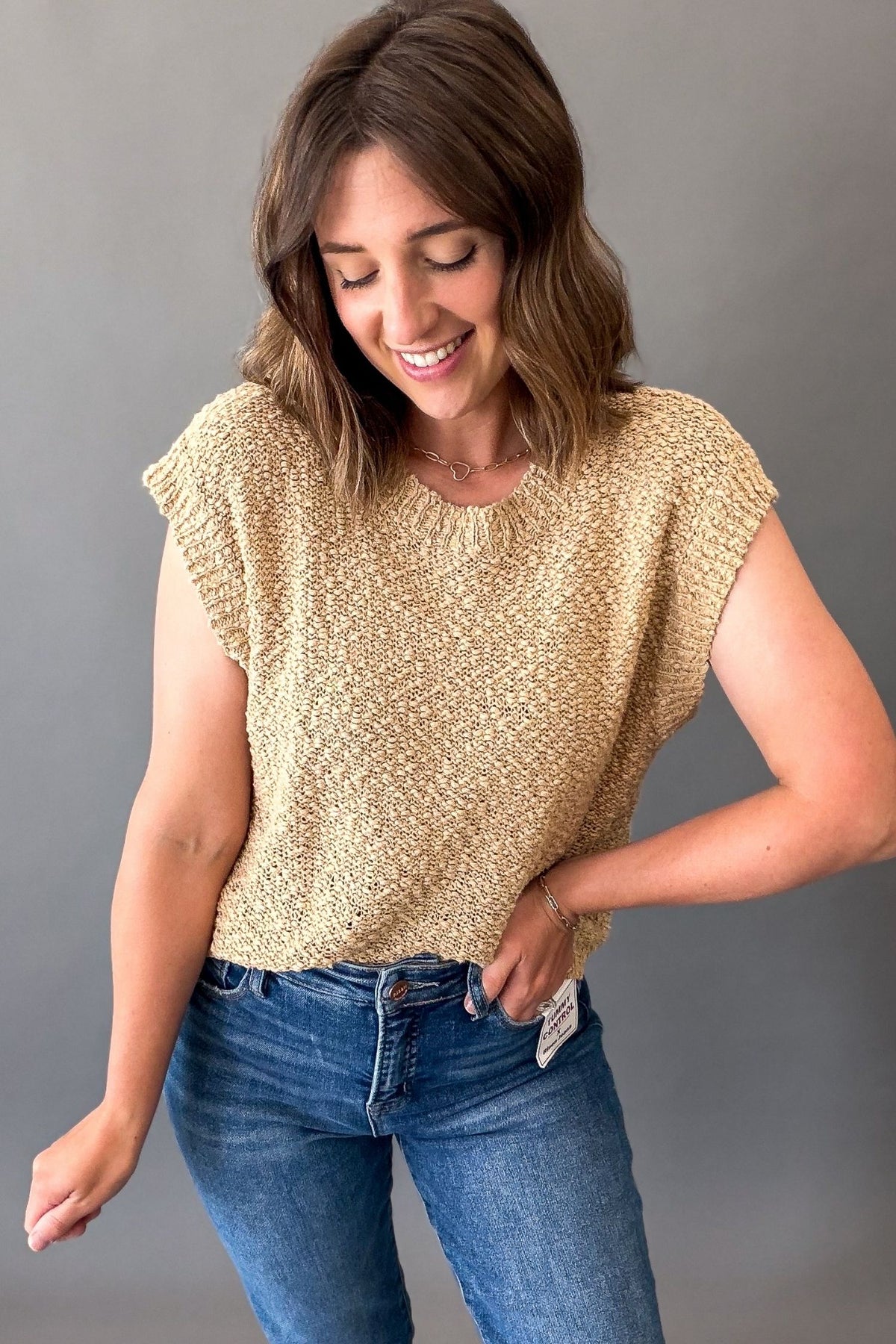 A woman wearing a textured, short-sleeve knit top in a warm beige or light tan color, paired with blue jeans, posing against a solid gray studio background.