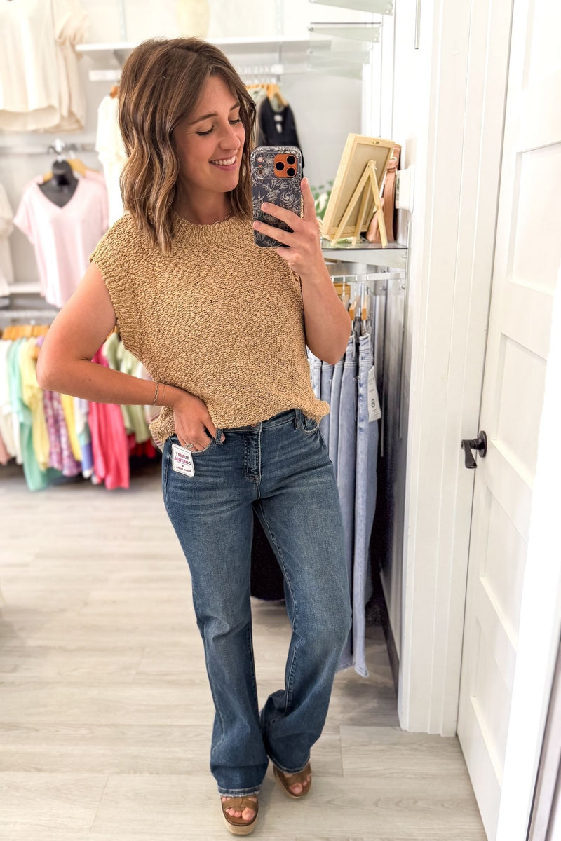 A woman takes a mirror selfie in a clothing boutique, wearing a textured biscuit colored sleeveless knit top and blue flare jeans.