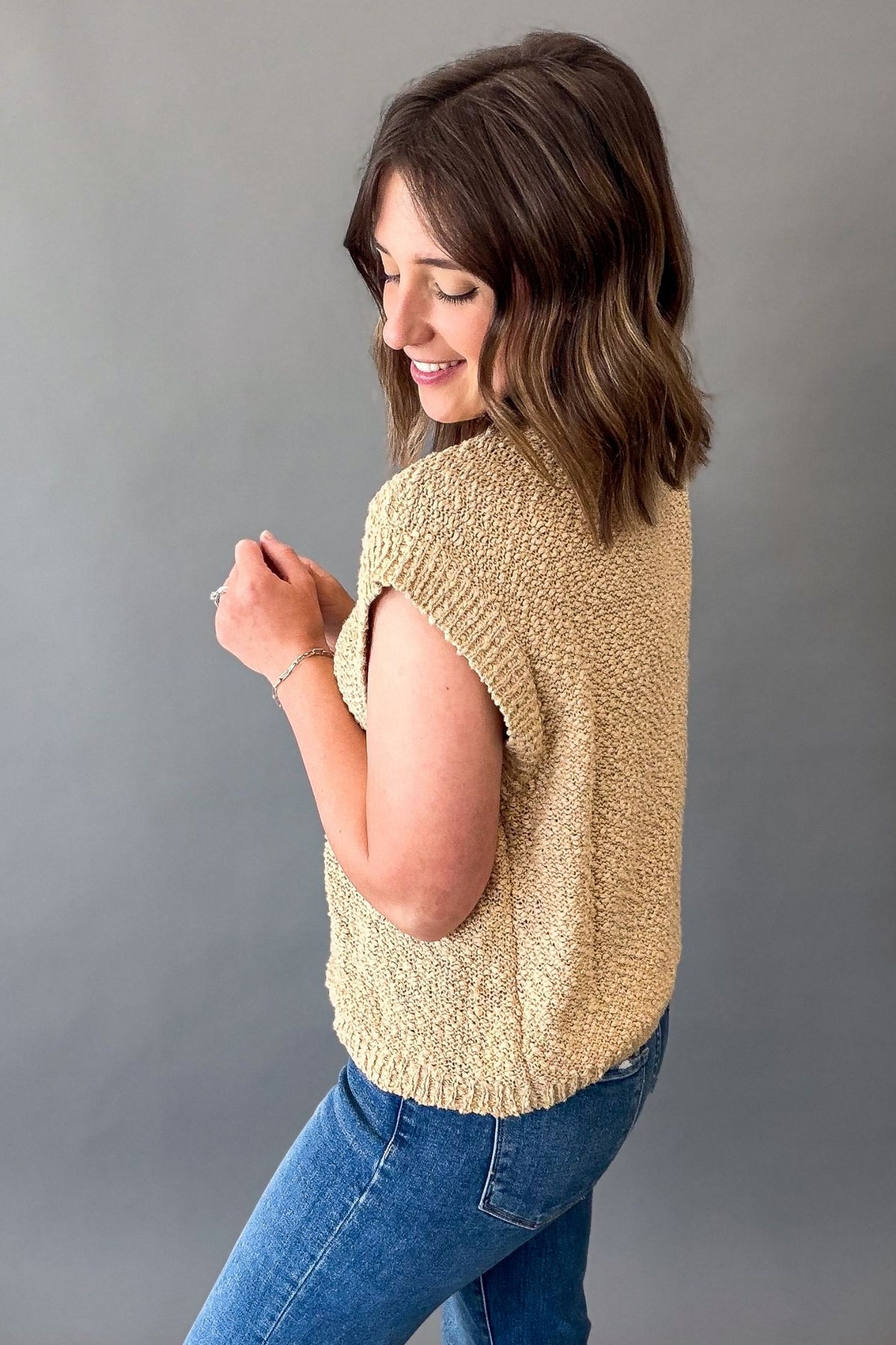 A woman wearing a textured, sleeveless knit top in a biscuit color, paired with blue jeans, posing against a solid gray studio background.