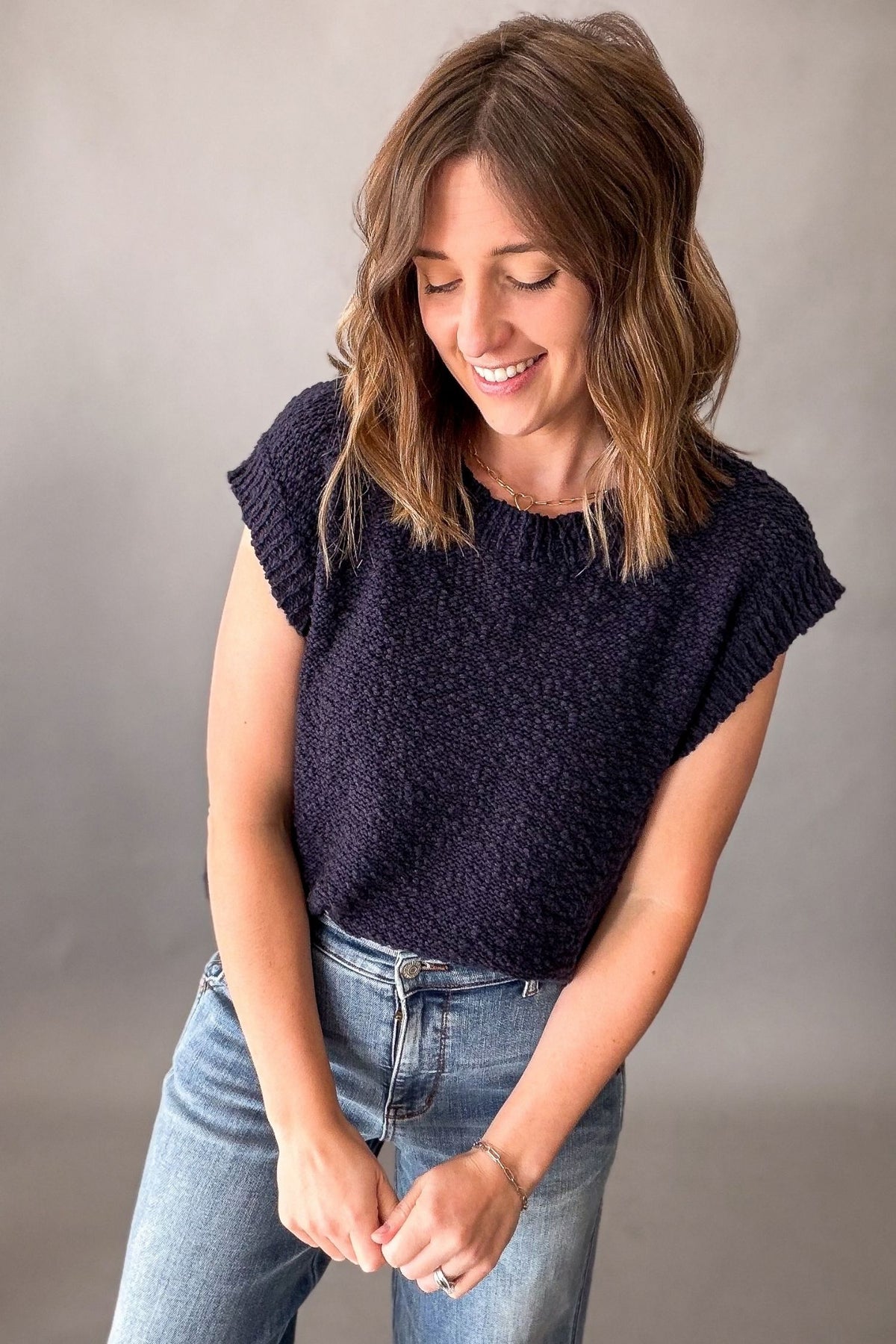 A woman wearing a dark navy textured knit short-sleeve top and light blue jeans, standing against a neutral gray studio background.