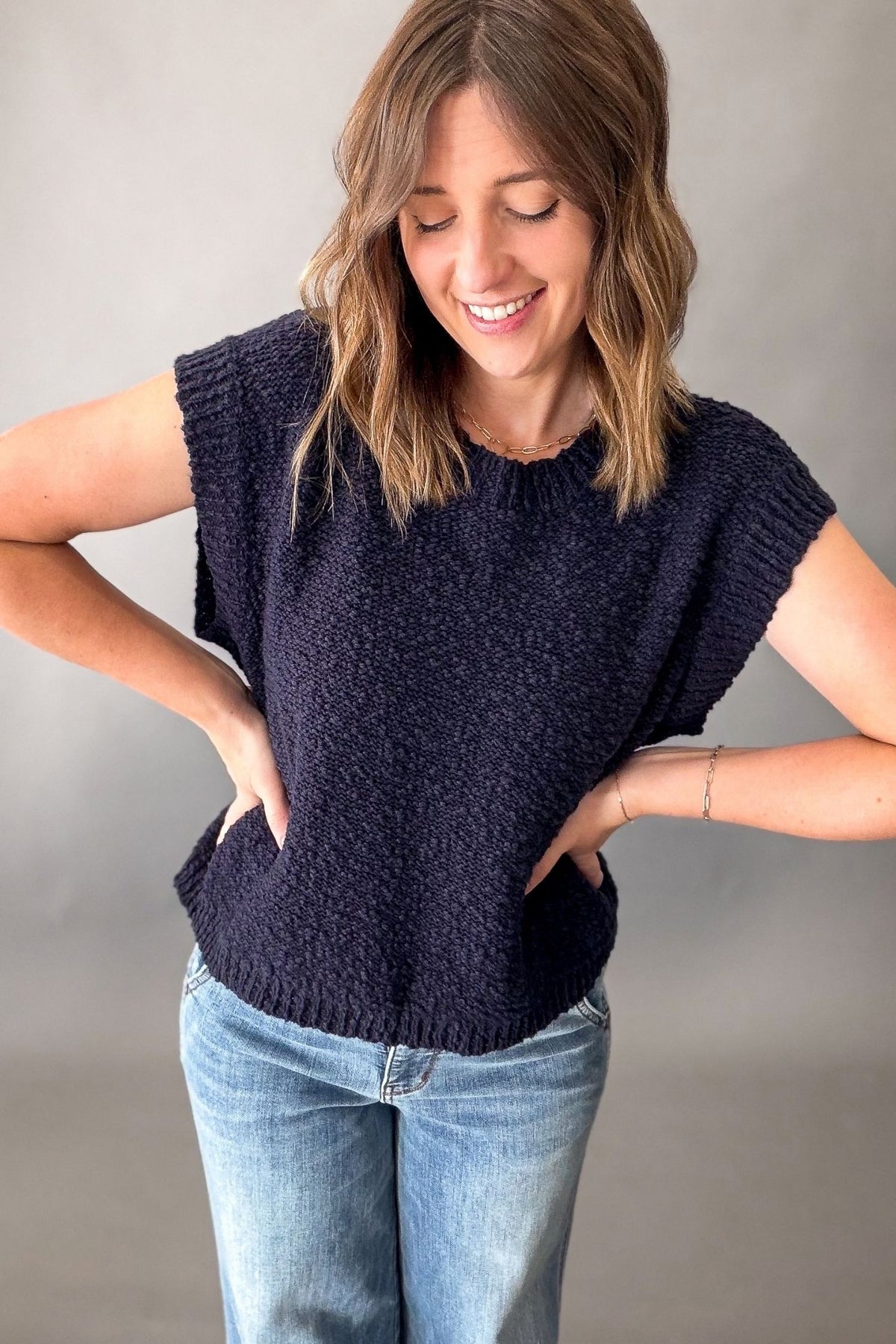 A woman wearing a dark navy blue textured knit sleeveless top and light blue jeans, posing with her hands on her hips against a neutral gray background.