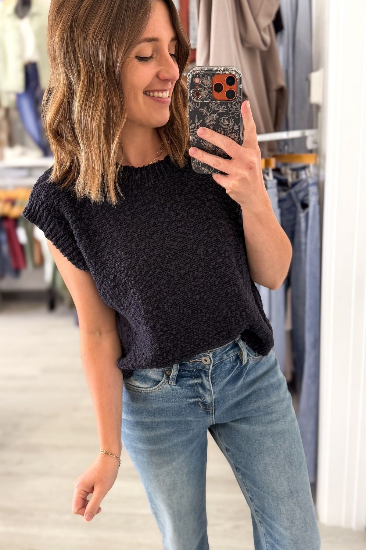 A woman takes a mirror selfie in a clothing store, wearing a dark charcoal textured short-sleeve top and light blue jeans.