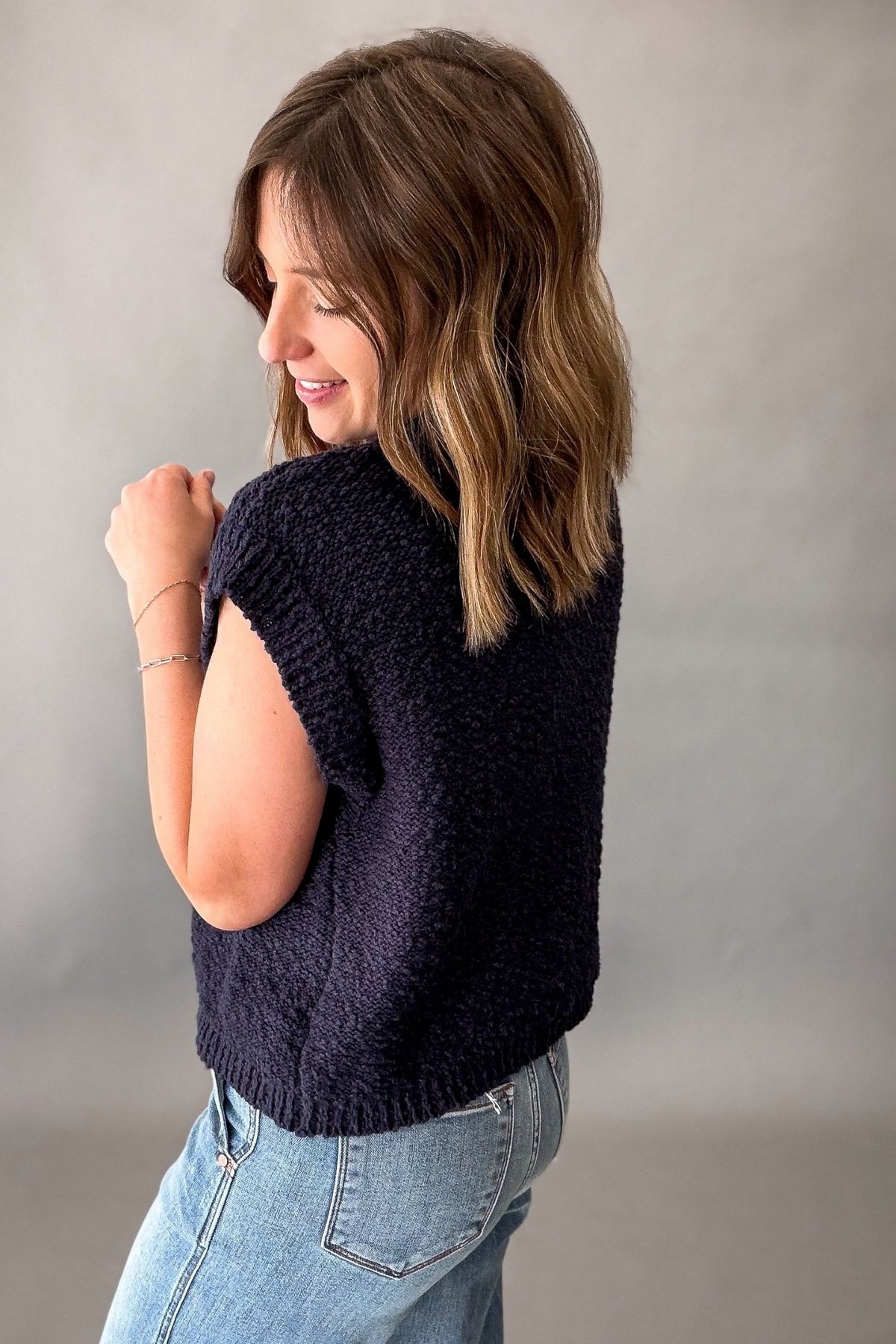A woman with shoulder-length brown hair is shown from the back wearing a dark navy textured knit sweater vest and light blue jeans, standing against a neutral gray studio background.