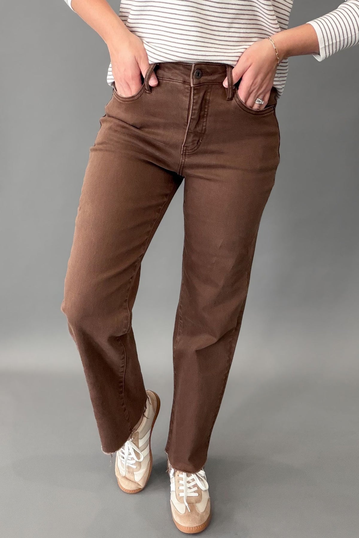 A person wearing brown straight-leg jeans, a white and mocha striped long-sleeve shirt, and beige sneakers, standing against a solid gray studio background.