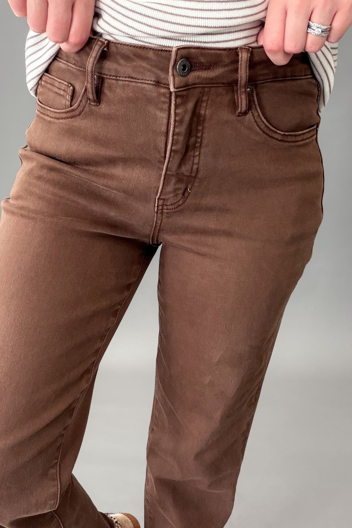A close-up of a person wearing brown straight-leg jeans, pulling the hem slightly to show the stretch and fit. The person is also wearing a white and mocha striped long-sleeve top, and a silver ring is visible on their finger, against a neutral gray studio background.