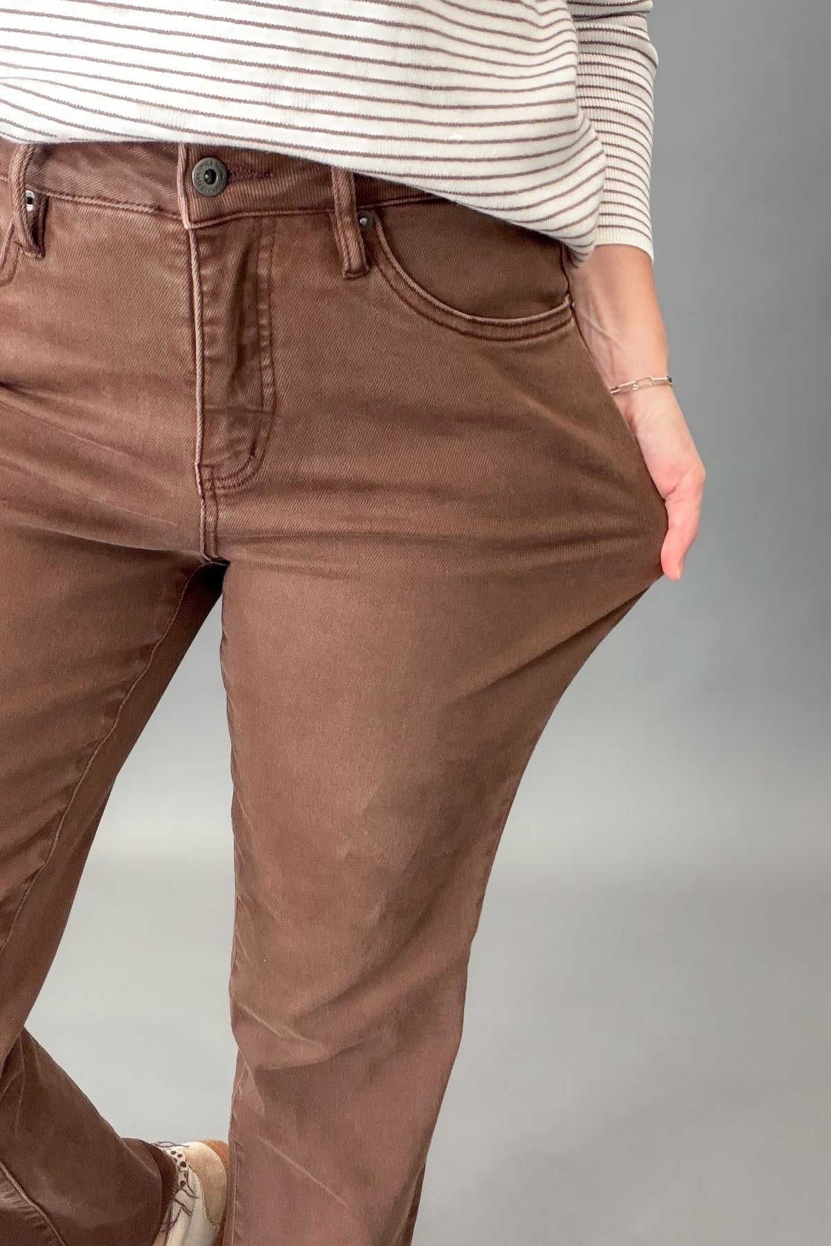 A close-up of a person wearing brown ankle straight jeans, pulling the fabric outward to demonstrate the stretch. The person is also wearing a cream and brown striped long-sleeve shirt, and the background is a solid gray studio backdrop.