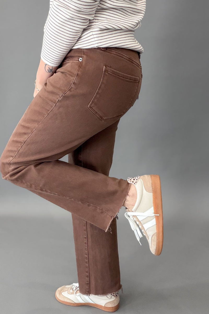 A person wearing brown straight-leg jeans, a striped long-sleeve shirt, and white sneakers with gum soles, shown from the waist down against a plain gray studio background.