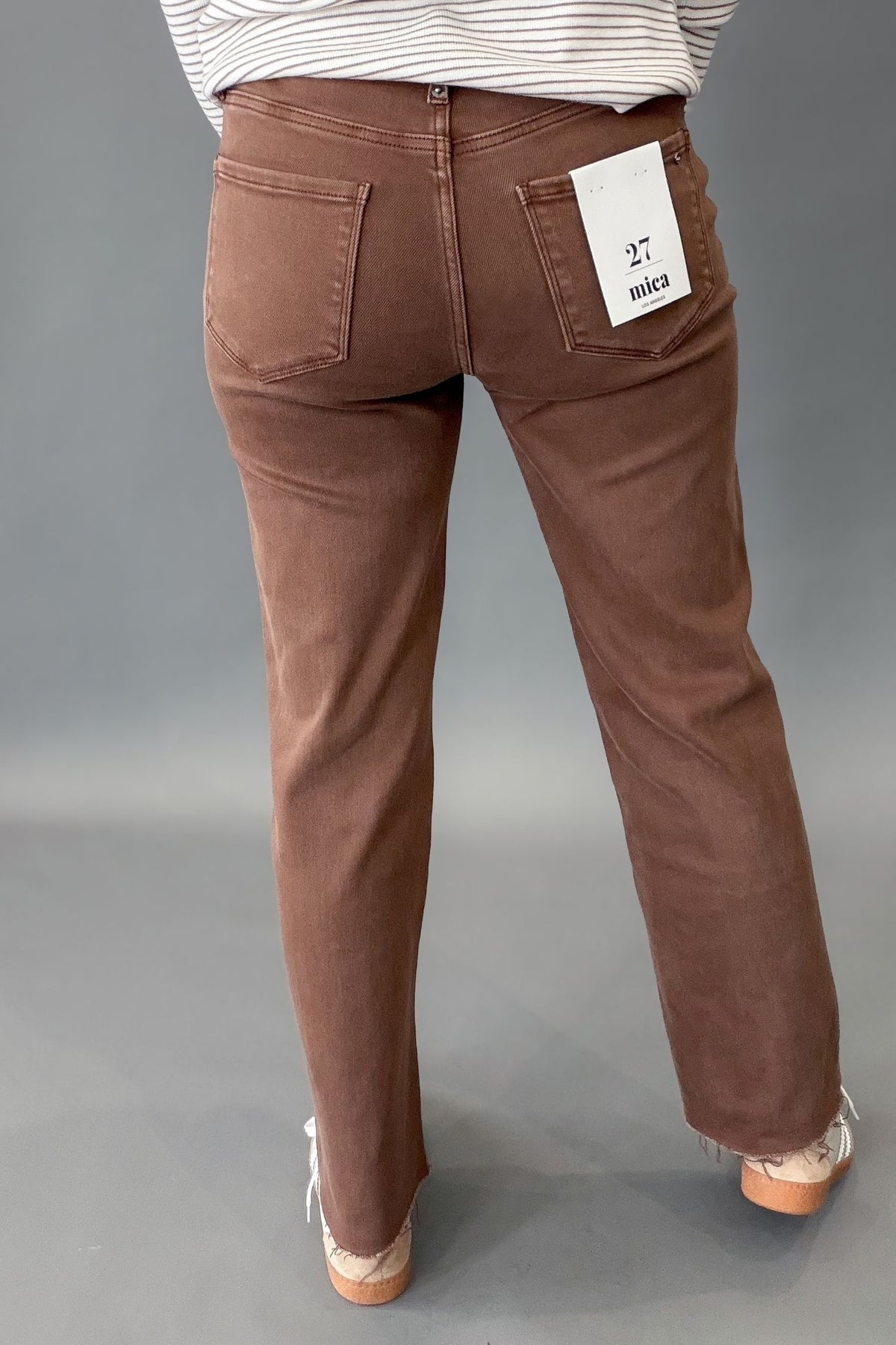 A person shown from the back wearing brown straight-leg jeans with a white size tag on the back pocket, paired with a white and mocha striped top and white sneakers, against a gray studio background.