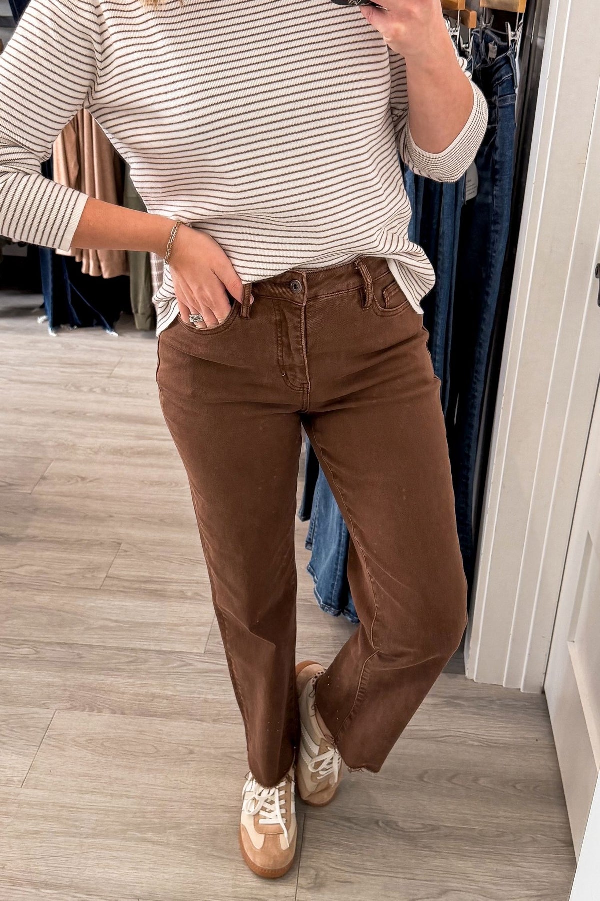 A person wearing brown straight-leg brown pants, a mocha and white striped long-sleeve shirt, and beige sneakers, standing in a clothing store.